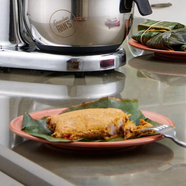 A cooked, opened Puerto Rican pastel on a plate next to Grandma Ann's Electric Grater and a stack of wrapped pasteles