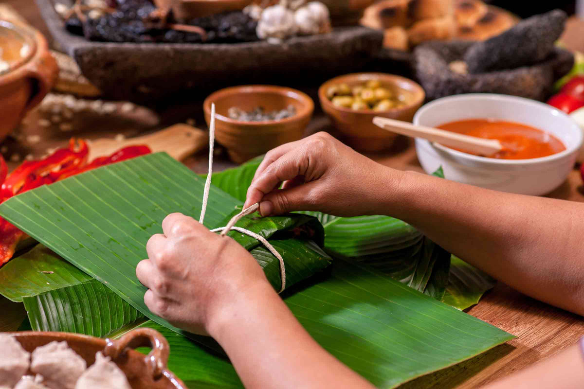 Hands tying a wrapped Puerto Rican pastel in banana leaves, surrounded by traditional ingredients
