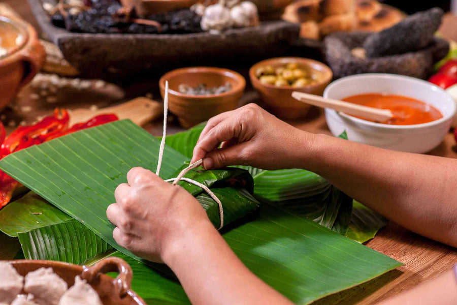 Hands tying a wrapped Puerto Rican pastel in banana leaves, surrounded by traditional ingredients