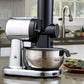 A countertop kitchen scene showing a black and silver electric grater with a stainless steel bowl, with a plate of puerto rican pasteles in the foreground 