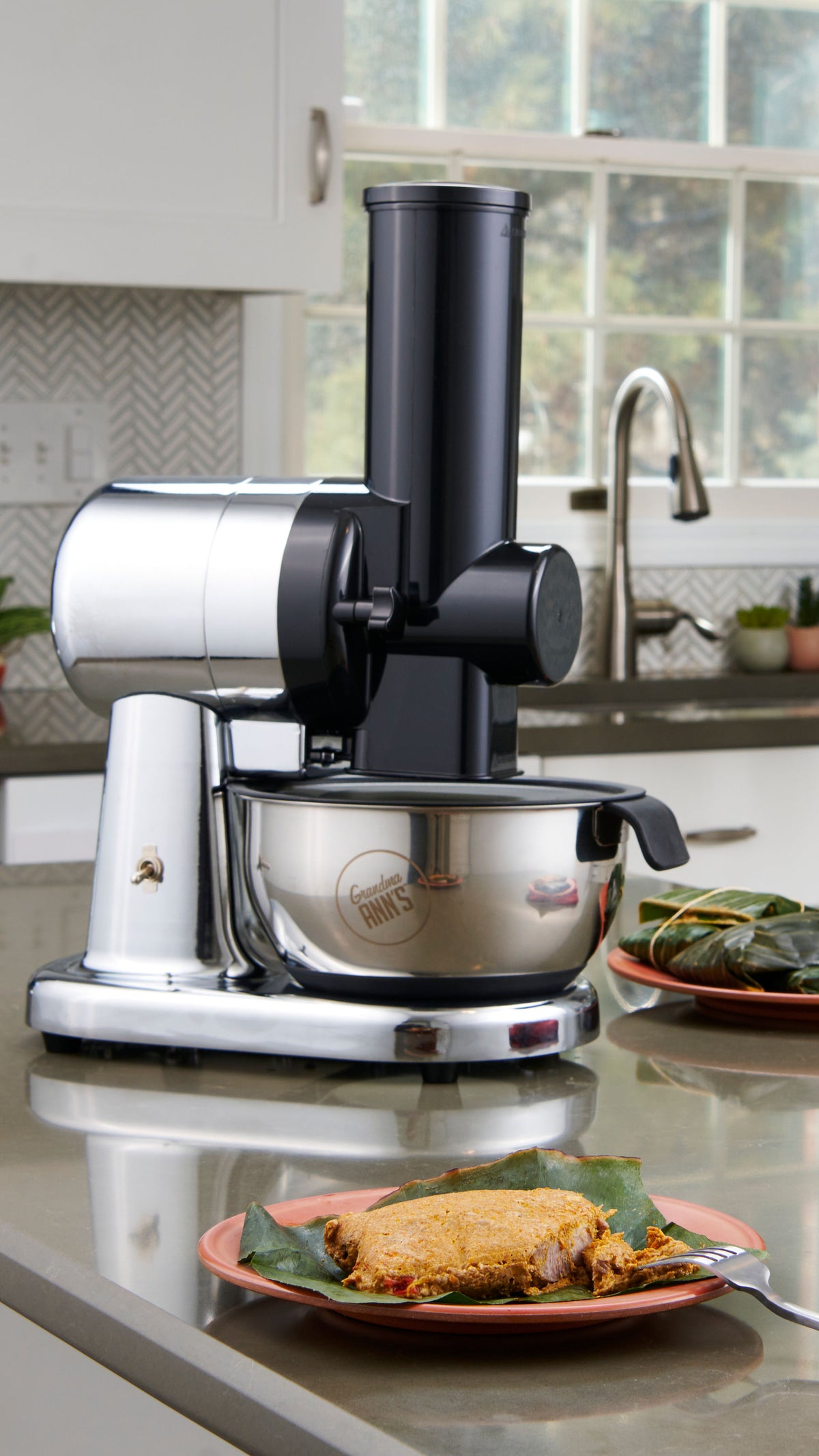 A countertop kitchen scene showing a black and silver electric grater with a stainless steel bowl, with a plate of puerto rican pasteles in the foreground 