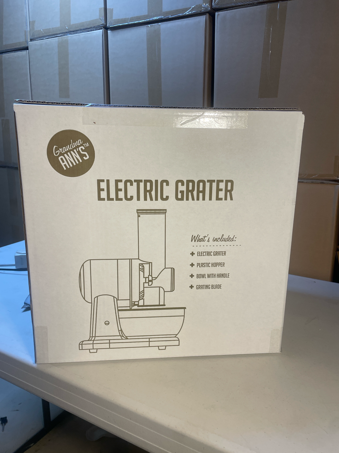 La Ralladora Eléctrica de Grandma Ann's en su empaque de venta, con cajas de inventario apiladas detrás
